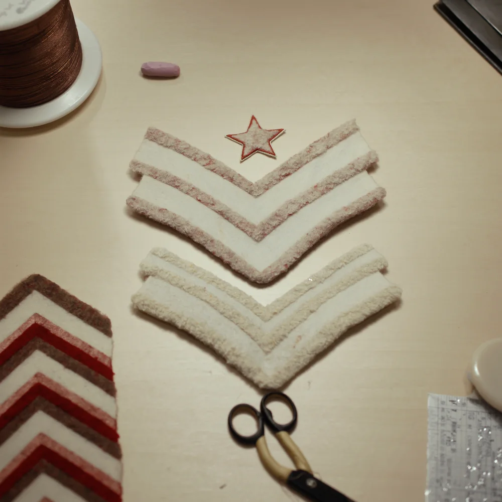 Blank felt chevrons, an embroidered star, a spool of gold thread, tailor's chalk and heavy shears laid out on a pale work table.