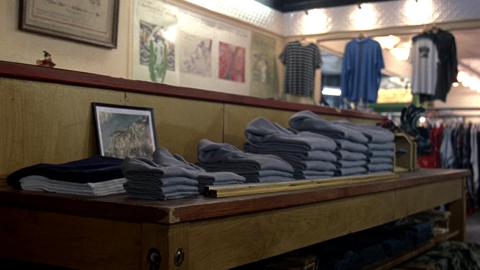 Folded stacks of plain sweatshirts and tee shirts on a long oak table in an old clothing store, framed prints on the wall behind.