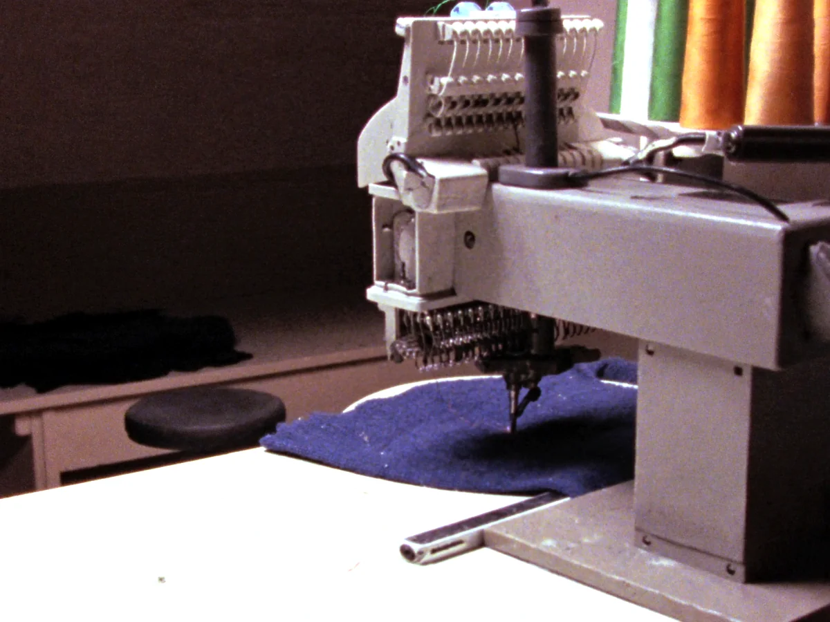 A single head commercial embroidery machine part way through a run, hooped fleece under the needle head with cones of thread on the rack above.
